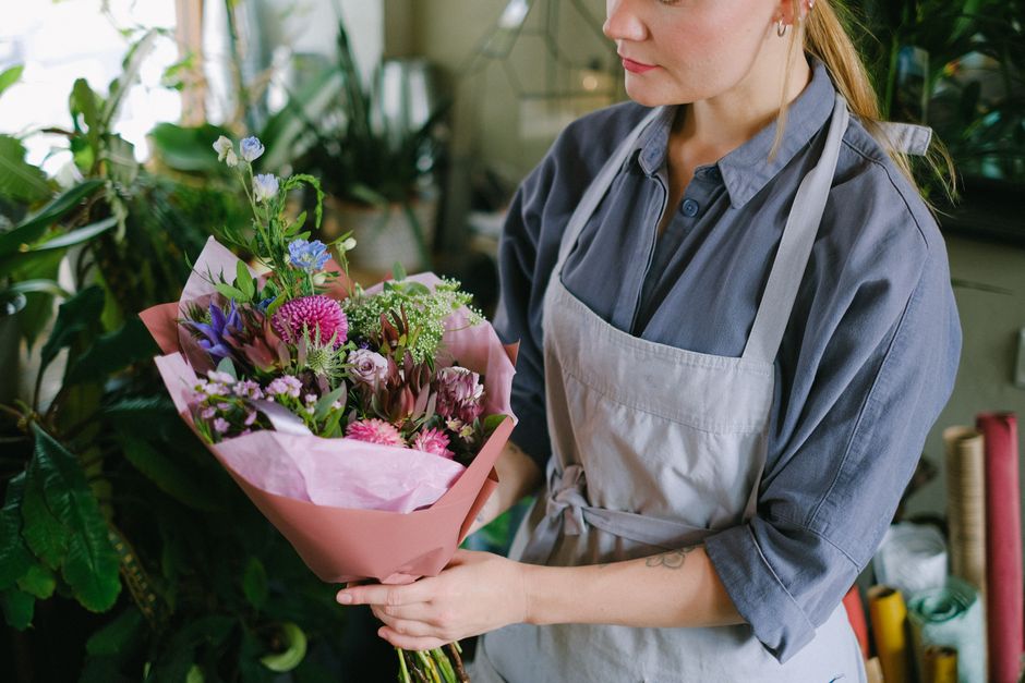 A woman wearing a grey shirt and beige apron holding a vibrant bouquet of fresh flowers wrapped in pink paper, with pink, purple, and white blossoms, including roses, carnations, and greenery, in an i