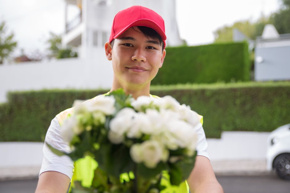 A friendly flower delivery person wearing a red cap and high-visibility vest is smiling and holding a bouquet of fresh white flowers, possibly roses or ranunculus, wrapped in clear cellophane. The bou