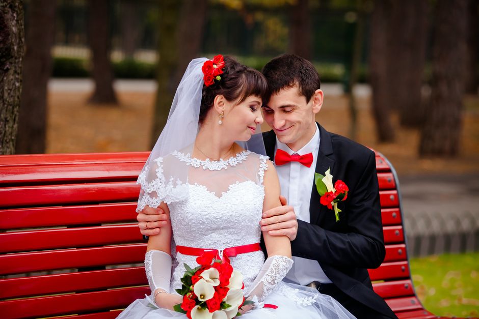 A bride and groom sitting closely on a red park bench during daylight, with the bride in a white lace wedding dress featuring a sheer neckline and floral embellishments, wearing elbow-length lace glov