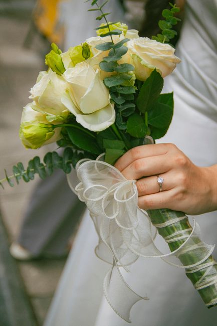 A close-up of a bridal bouquet held by a bride, featuring creamy white roses and delicate green buds, accented with sprigs of eucalyptus and other greenery. The flowers are fresh with soft, velvety pe