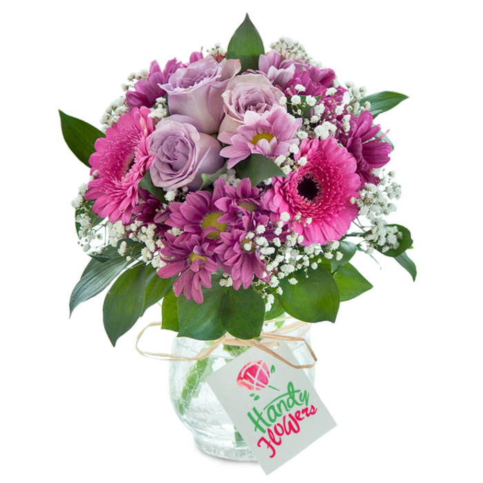 Elegantly abundant yet intimate, this mixed bouquet is thoughtfully conceived to reflect the charm and character of Forest Hill's vibrant local life. At its centre, a cluster of soft lavender roses reveal their gently curled edges, their subtle fragrance recalling the gentle breeze through Sydenham Woods. Encased by striking fuchsia pink Gerbera daisies-whose wide, open petals and distinctive dark centres express contagious cheer-the composition is further enriched by clusters of deep magenta chrysanthemums, their plum-toned blooms radiating fresh energy and natural texture. Tracing the arrangement are airy sprays of white baby's breath, imparting a dreamy lightness and creating visual space among the richly saturated petals. Framing the vivid display, broad, glossy green leaves bring a grounding touch, reminiscent of the sweeping greenery around Horniman Museum. The entire arrangement gathers in a dense, well-rounded form inside a clear glass vase-its finely cracked surface offering a hint of rustic elegance, as if crafted from the textures and light unique to Forest Hill's artisan shops. A strand of natural raffia is tied around the neck, suspending a crisp white gift tag festooned with lively birthday lettering and a sweet floral accent, capturing the spirit of local festivity. This bouquet, with its layered shades and tactile intrigue, is an inspired choice for any special occasion in SE23-a floral tribute as welcoming and expressive as Forest Hill itself.