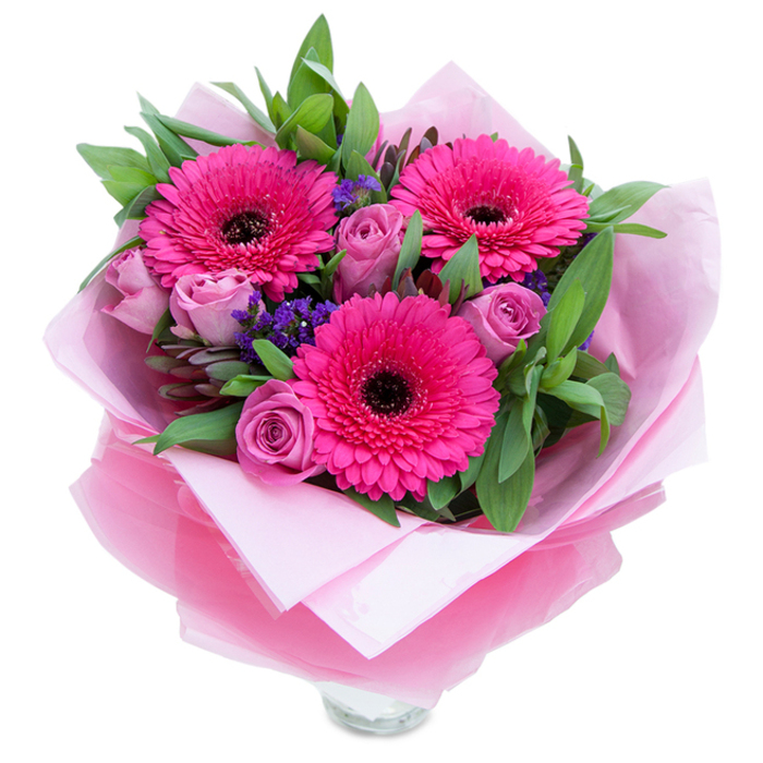 I see this bouquet as a balance of boldness and tenderness: three fuchsia Gerbera daisies stand proudly, their vivid petals radiating from dark, almost black centres that give the arrangement focus and energy. Close to them, lavender-pink roses open in gentle spirals, their velvety, dusty-rose tones providing a softer, more private rhythm. Tiny sprays of purple statice are tucked throughout, offering a papery texture and small, jewel-like flashes of cool colour that punctuate the larger shapes. Broad green leaves - the kind you'd expect from salal or eucalyptus - frame the blooms, adding weight and a fresh, garden-scented backdrop. The bouquet is wrapped in an inner layer of matte pale pink tissue and an outer veil of translucent fuchsia-pink cellophane with a faint crinkle that catches the light; the wrapping is folded with intention, presenting the stems neatly above the visible rim of a clear glass vase. Under diffused daylight the pinks read both warm and lively, while the overall effect feels celebratory yet intimate. The fragrance profile is subtle: a whisper of rose perfume with the understated green note of the Gerberas and foliage. Picture this piece brightening a breakfast table on Perry Vale, arriving at a small gathering near Forest Hill Library, or resting on a bedside table as a thoughtful pick-me-up - a versatile, cheerful choice for birthdays, anniversaries, or simply to say 'you matter'.